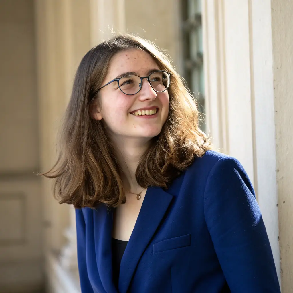 Jane has long blonde hair and blue eyes, and she is wearing glasses and a black turtleneck.