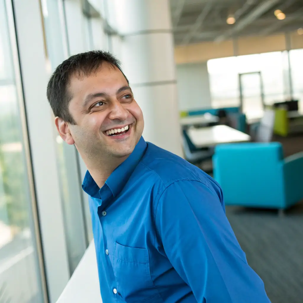 A man with dark hair, wearing a blue shirt and a friendly smile, standing in a modern office.