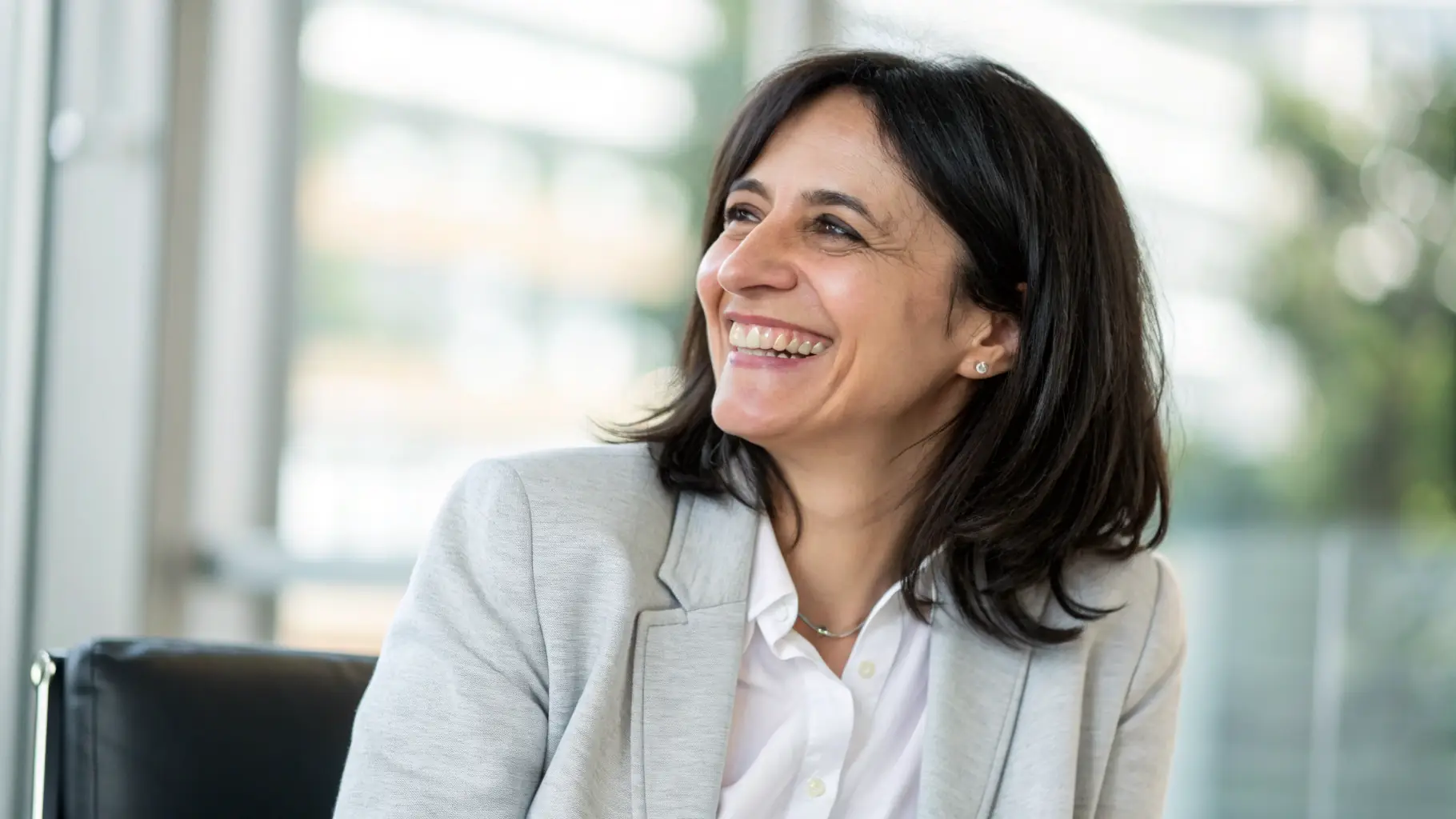A person is smiling and talking to the camera in a well-lit office setting. The background is blurred to focus on the speaker.