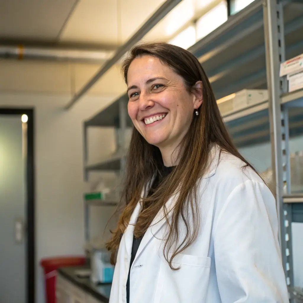 A smiling woman with short brown hair and glasses, wearing a white lab coat.