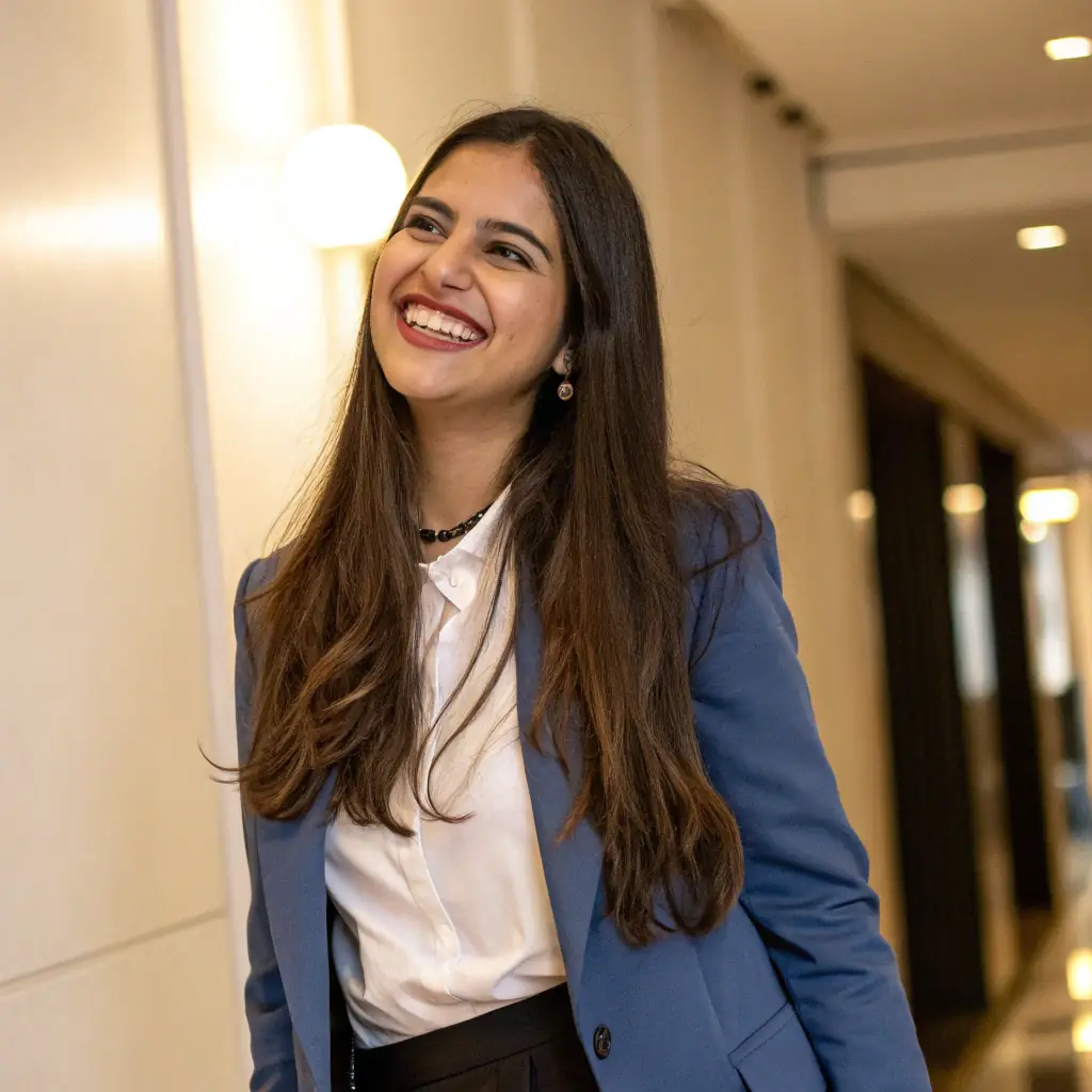 Aisha is a young woman with long brown hair and a bright, engaging smile. She wears a colorful scarf and a professional blouse.
